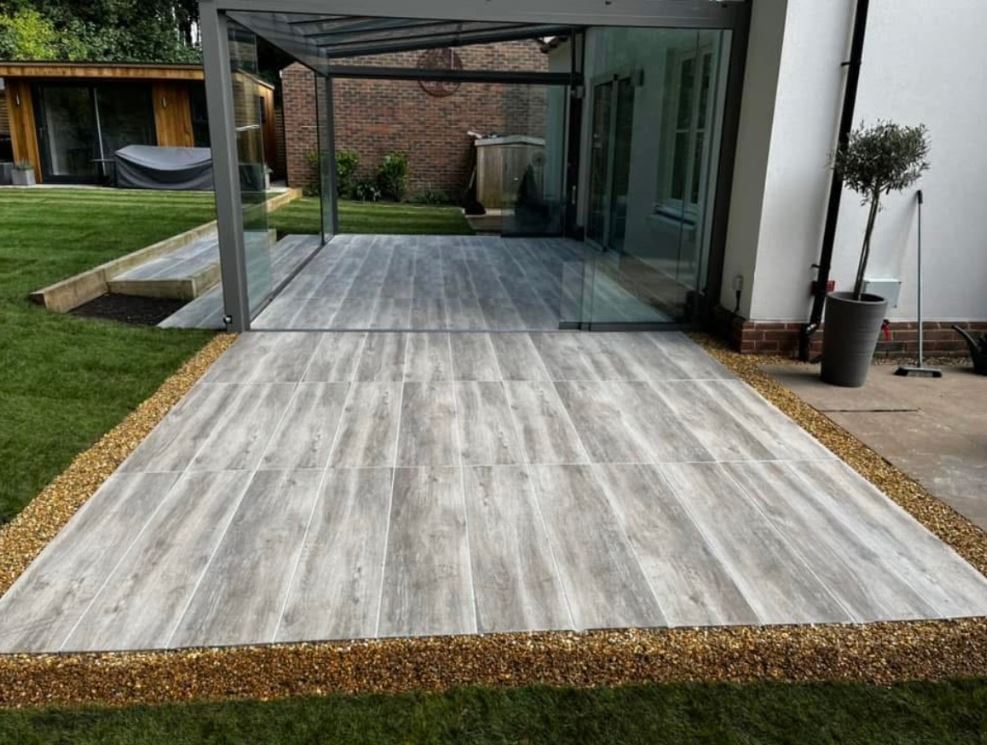 A grey porcelain-plank terrace with a gravel border, running out from a glass veranda to a level lawn.